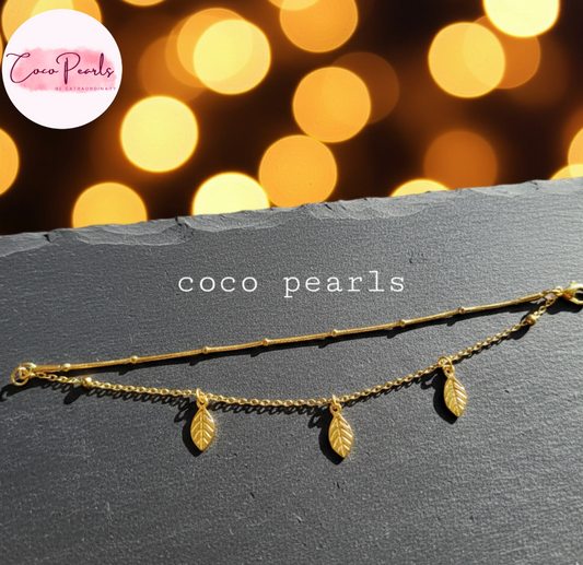 coco pearls stainless steel anti tarnish Gold leafy whispers bracelet with leaf pendants on a dark surface, blurred lights in the background,
