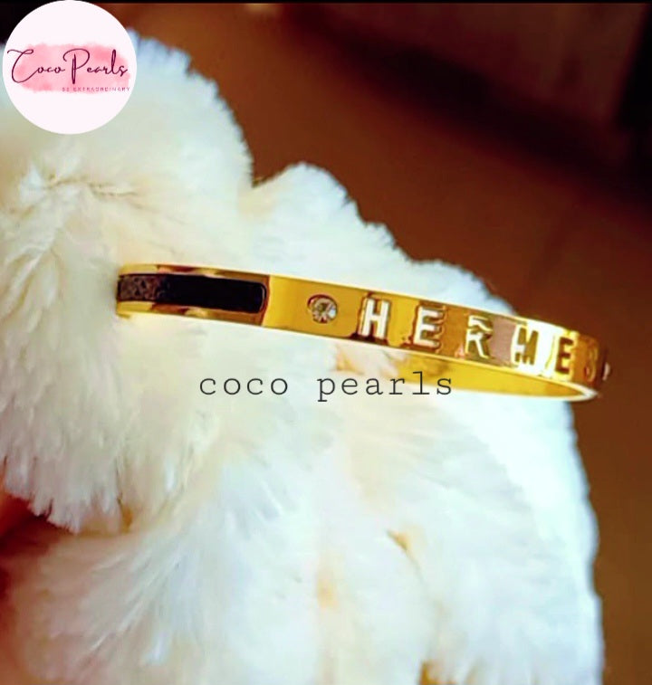 Coco Pearls Stainless Steel Anti Tarnish Gold  lunar glow handcuff bangle bracelet with 'HERMES' engraving on a white fluffy surface