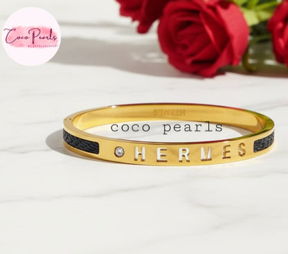 Coco Pearls Stainless Steel Anti Tarnish Gold  lunar glow handcuff bangle bracelet with 'HERMES' engraving on a white surface with red roses in the background.