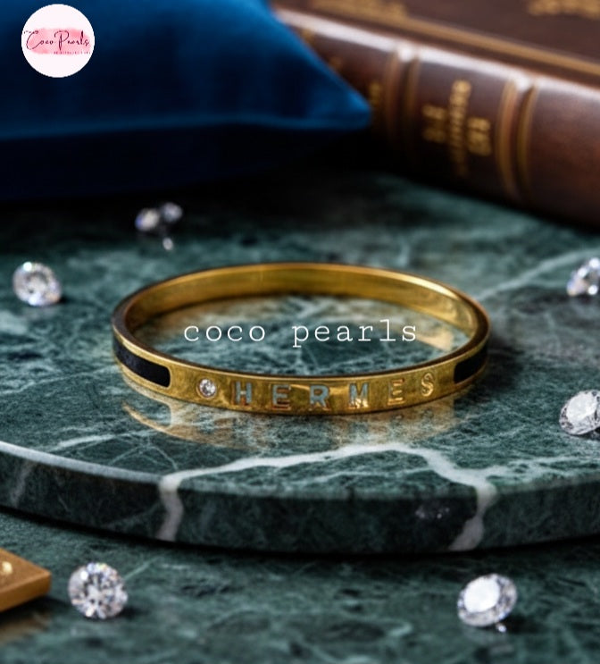 Coco Pearls Stainless Steel Anti Tarnish Gold  lunar glow handcuff bangle bracelet with 'HERMES' engraving on a green marble surface