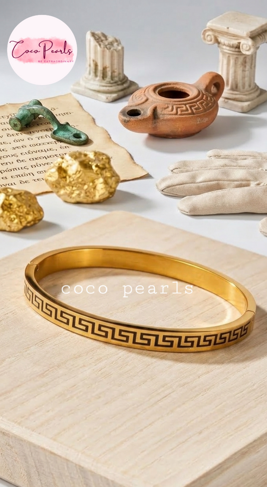 Gold bracelet with Greek key design on a light wooden surface, with 'Coco Pearls' branding.