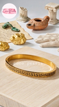 Gold bracelet with Greek key design on a light wooden surface, with 'Coco Pearls' branding.