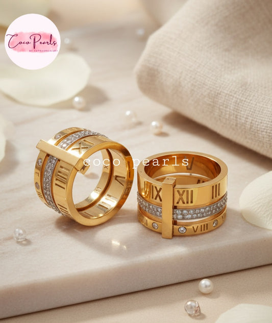 Coco Pearls Stainless Steel Anti Tarnish Two gold roman rings with Roman numeral design, diamond band and numeral diamond band on a soft beige background'.