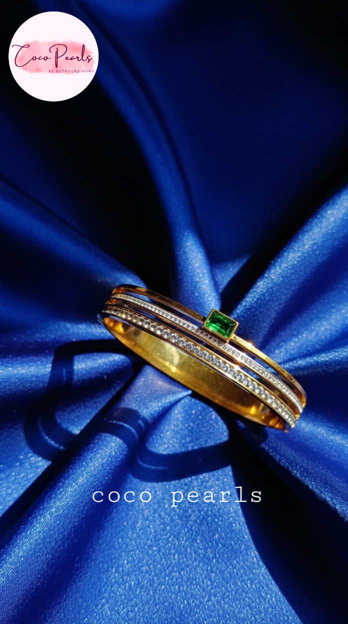 coco pearls stainless steel anti tarnish gold royal emerald diamond handcuff bangle bracelet with rhinestones and emerald in center displayed on a blue background