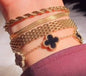 Coco Pearls Stainless Steel Anti Tarnish four Gold timeless charm bracelet stack with a black clover charm bracelet, mesh bracelet, snake chain bracelet and rope bracelet on a wrist against a blurred background