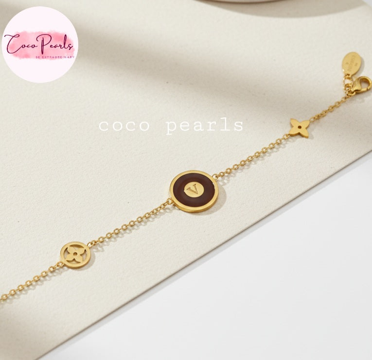 Coco Pearls Stainless Steel Anti tarnish Gold and brown v link bracelet with v initial on brown circular charm in center and golden charms on a beige surface