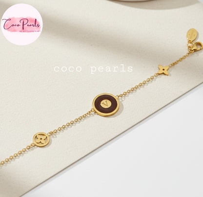 Coco Pearls Stainless Steel Anti tarnish Gold and brown v link bracelet with v initial on brown circular charm in center and golden charms on a beige surface